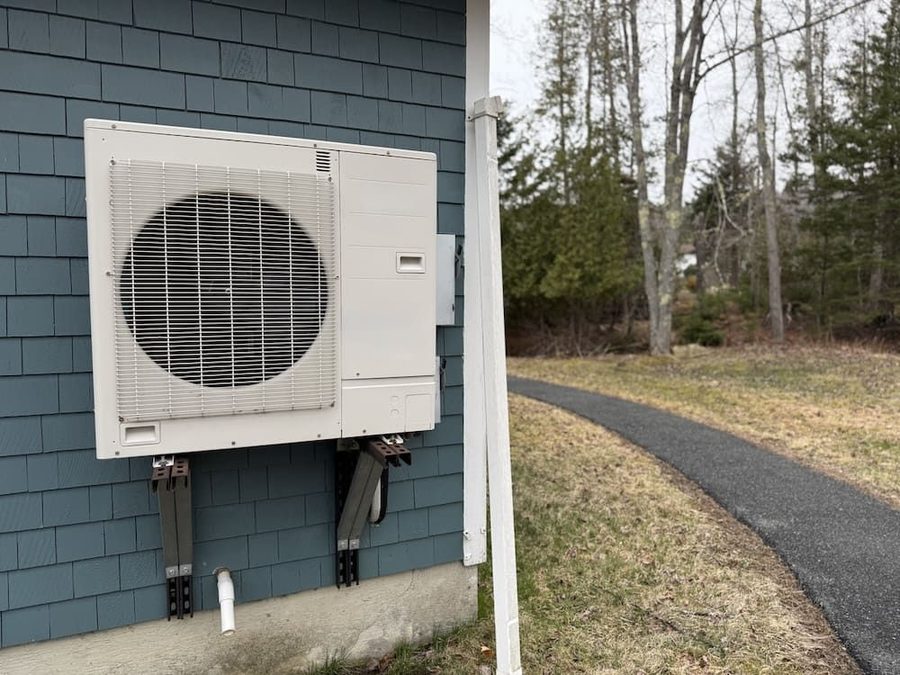 Heat pump on the side of a house; there is a black concrete pathway along the side of the house.